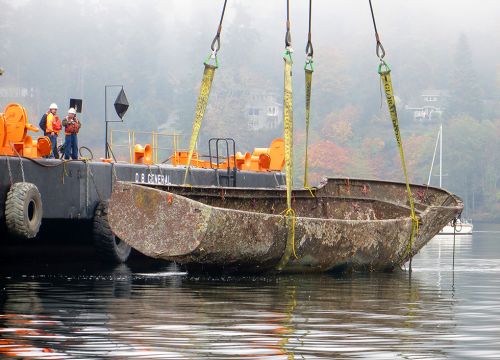 Workers direct the lifting of a rusted boat from a waterway onto a barge.