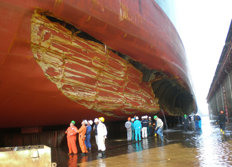 People observe a large tanker with a huge gash in its hull in dry dock.