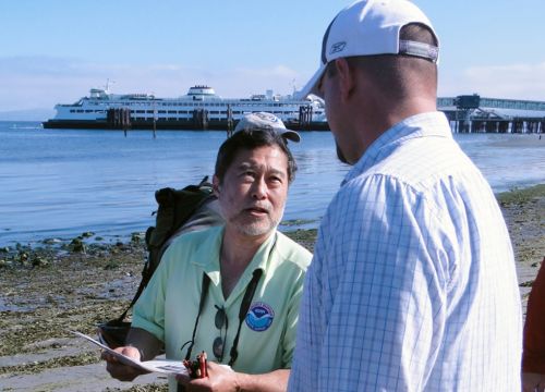 A NOAA spill responder with a clip board talking on a beach with a ferry behind.