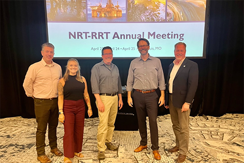 Five OR&R team members pose in a group photo in front of a presentation screen with a title slide reading "NRT-RRT" Annual Meeting.