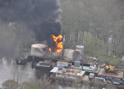 Fire burns in one of several oil tanks on a platform in a bayou.