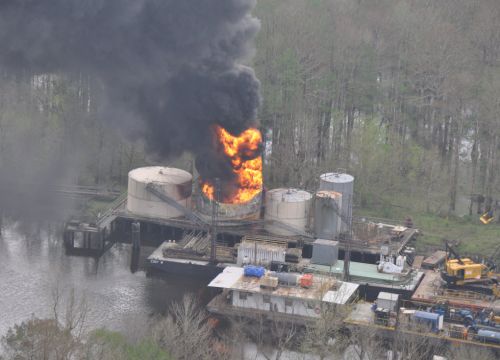 Flames and dark smoke billow from oil production platform fire next to a bayou. 