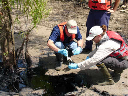 Two spill responders scoop oil into a sampling jar at an oiled shoreline.