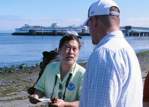 Instructor talks with student on a beach, with ferry in background.