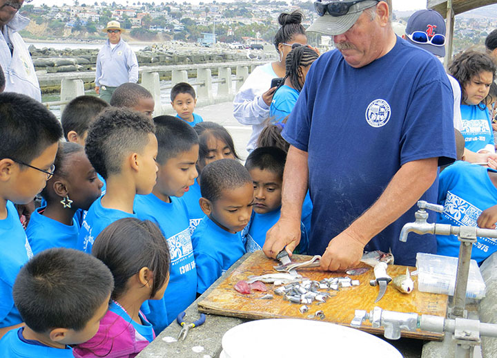 A group of kids surround a man filleting fish on a pier.