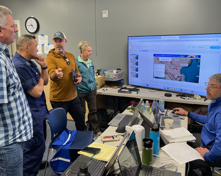 A group of individuals talk together in front of a monitor displaying a presentation of oil spill briefing materials.