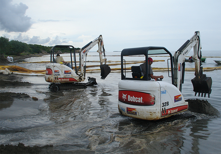 Two bobcat digging machines scoop oil from a beach.
