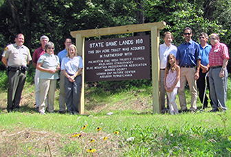 Groups of people standing by a state game lands sign in front of woods.