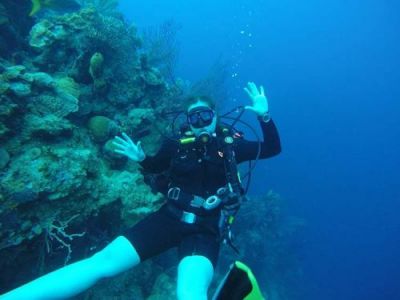 A woman in SCUBA diving gear smiles underwater next to a coral reef.