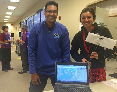 A man and a woman stand behind an open laptop on a table while they pose for a photo. The woman is holding a small sign that says "GIS."