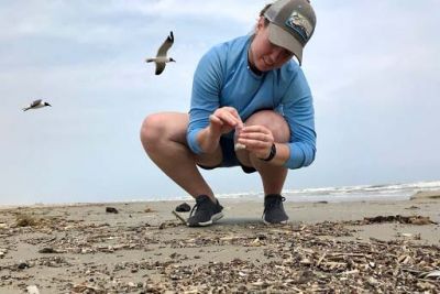 A woman bends down on a beach to put small plastics items into a plastic bag.