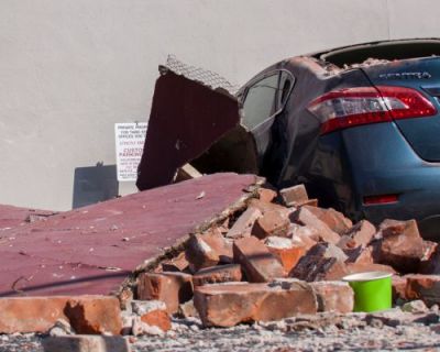 A car with a for sale sign heavily damaged by falling bricks and other debris during an earthquake.