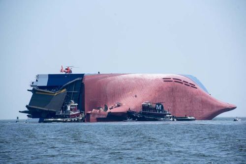 A large freighter vessel overturned on its site with a helicopter on it and several smaller vessels surrounding it.