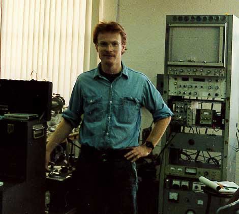 Man standing with computer equipment in the background.