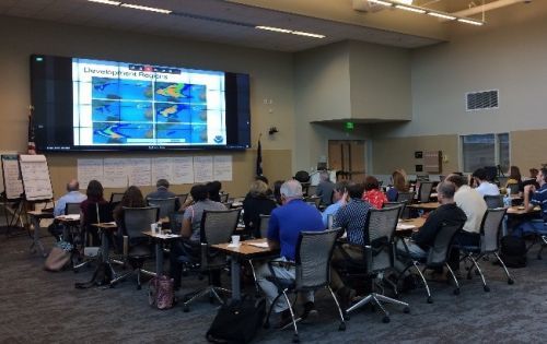 Adult students in a large classroom setting look up at a large TV monitor.
