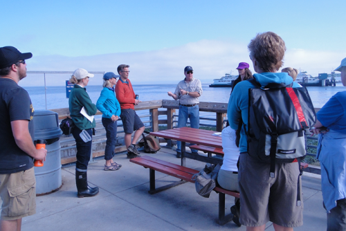 A group of people standing outside in front of a body of water with vessel traffic. 