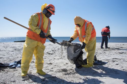 Two people in hazmat suits clean up oil from a beach.