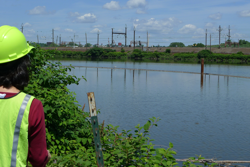 A person in a hard hat looking across a river at an industrial shoreline.