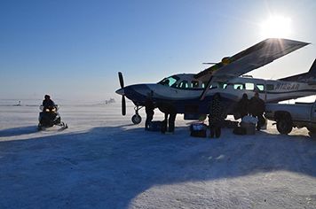 People load a propeller plane while a person approaches on snowmobile.