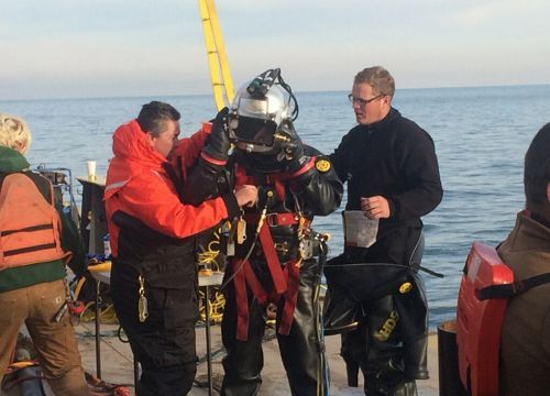 Two people on a boat inspect a diver in a full dive suit.