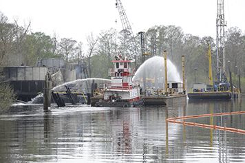 Fire-fighting vessel sprays water on an oil tank on a platform in a bayou.
