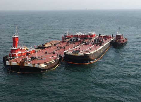 A large barge is being offloaded next to tugboats in the ocean.