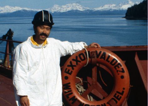 A man in a tyvek suit stands on a ship next to a life preserver in Alaska.