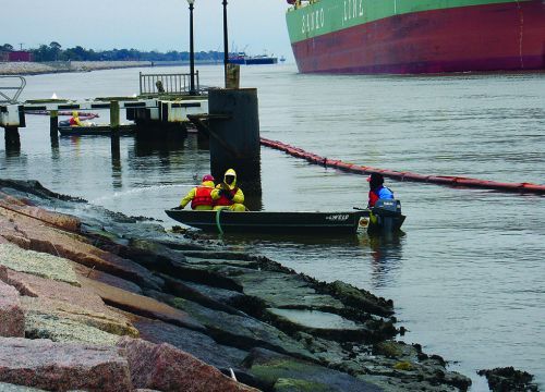 Responders in a small boat pressure-wash rocky shore at the site of an oil spill