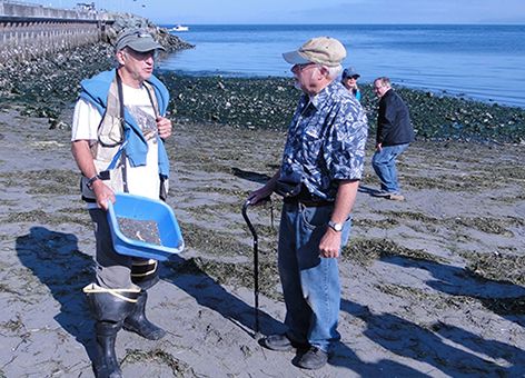 Two men on a beach with one holding a bin of sand.