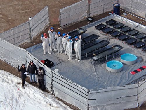 Aerial view of a group of oil spill specialists in PPE waving at the drone taking the shot. The group stands next to plastics totes staged during a field experiment in organized rows, each containing treatments of oil and diesel. The surrounding environment is icy.