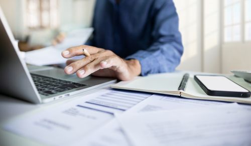 Man working online with laptop at office; documents and a notebook are seen to his left. Image credit: Lek via Adobe Stock.