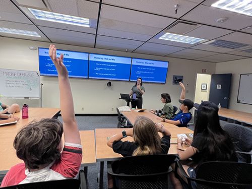 Students raise their hands to participate in a classroom setting activity led by a facilitator at the front of the classroom.