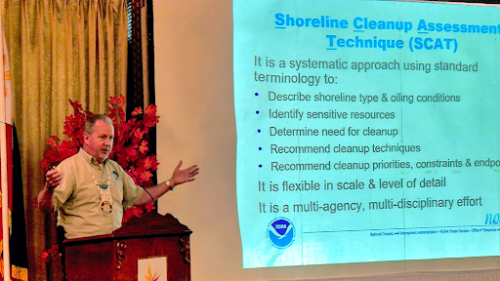 Individual presents in front of a room next to a projector screen that features information about the Shoreline Cleanup Assessment Technique.