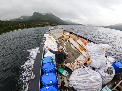 A person pointing to a mountainous shoreline while on a moving vessel that is full of various marine debris items.