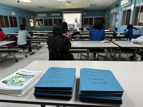 A presenter is speaking to a classroom of people with a stack of marine debris curriculum workbooks piled on one of the desks. 