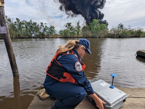 Responder sets up a box of monitoring equipment on a dock in the foreground. Large black smoke plume observed in the background of a marshland landscape.