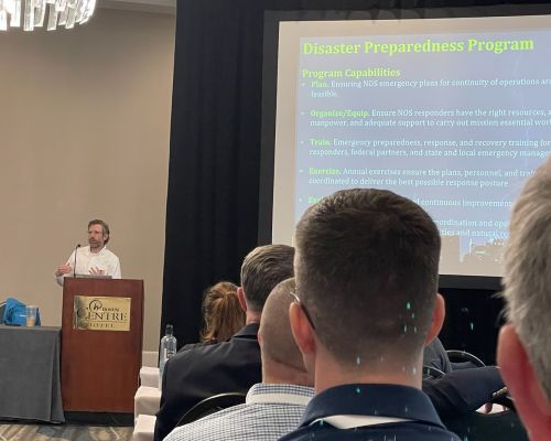 Man stands at a podium at the front of a conference room in front of a projection screen displaying a slide about the Disaster Preparedness Program.