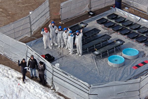 Aerial view of a group of oil spill specialists in PPE waving at the drone taking the shot. The group stands next to plastics totes staged during a field experiment in organized rows, each containing treatments of oil and diesel. The surrounding environment is icy.