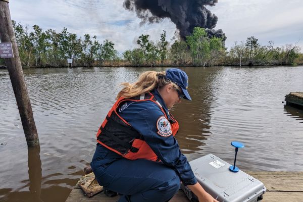 Responder sets up a box of monitoring equipment on a dock in the foreground. Large black smoke plume observed in the background of a marshland landscape.