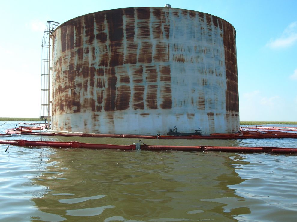Large oil tank surrounded by boom in water.