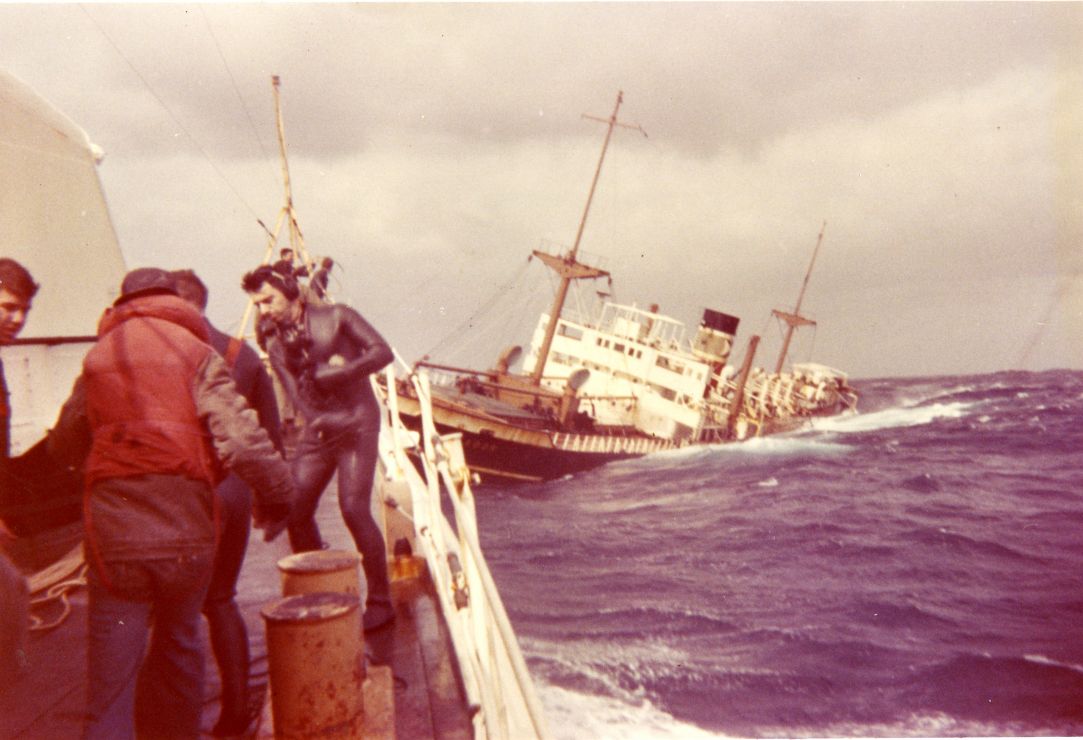 Responders, including one in a wetsuit, work on a vessel next to a sinking ship.