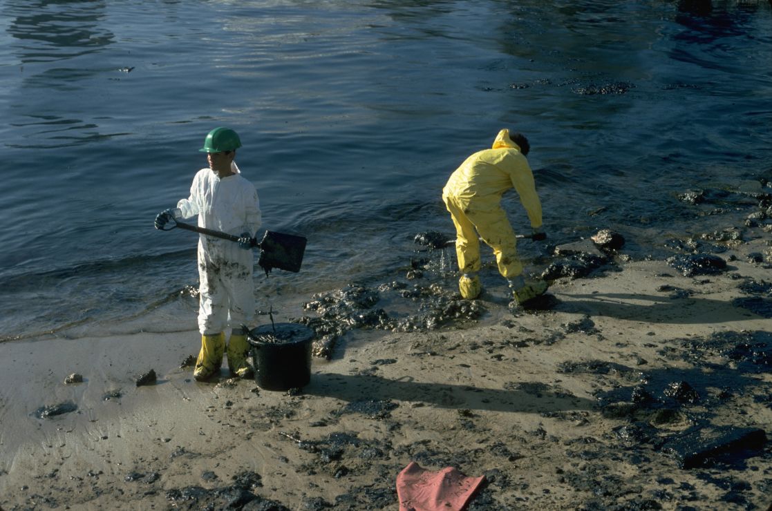 Photo of two workers shoveling black oil off of a beach.