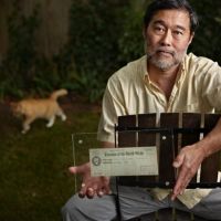 A man sitting on a chair holding a framed check with a cat in the background.
