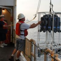 Two people launch a water column sampling device off the side of a ship.