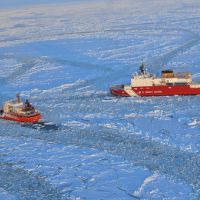 Coast Guard icebreaker breaks ice for a supply ship on an ice-covered ocean.