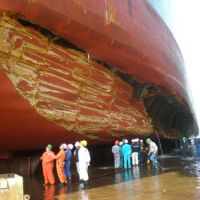 People observe a large tanker with a huge gash in its hull in dry dock.