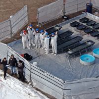 Aerial view of a group of oil spill specialists in PPE waving at the drone taking the shot. The group stands next to plastics totes staged during a field experiment in organized rows, each containing treatments of oil and diesel. The surrounding environment is icy. Image credit: CRRC.
