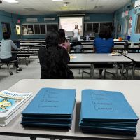 A presenter is speaking to a classroom of people with a stack of marine debris curriculum workbooks piled on one of the desks. 