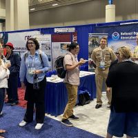 Conference attendees and booth representatives engaging in conversation within an exhibit hall.
