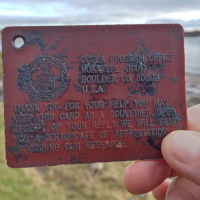A hand holding an old plastic drift card with reporting information imprinted on it held up in front of a shoreline.